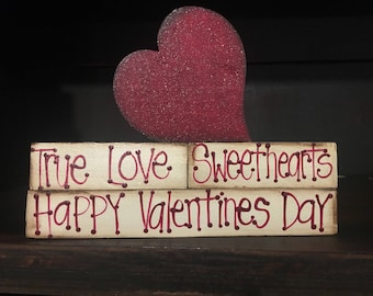May include: Three wooden blocks stacked on top of each other with the words "True Love Sweethearts Happy Valentines Day" written in red paint. A red heart cutout is placed on top of the blocks.