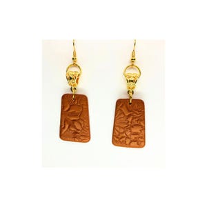 May include: A pair of earrings with a rectangular, brown pendant featuring a floral design. The earrings have gold-colored hooks and decorative accents. The pendants are approximately 1 inch in length.