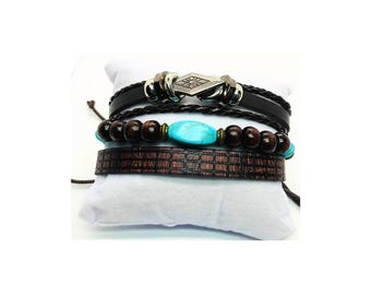 May include: A stack of four bracelets with a bohemian style. The bracelets include black leather, wooden beads, a turquoise stone, and a brown leather band with a brick pattern. The bracelets are displayed on a white pillow.