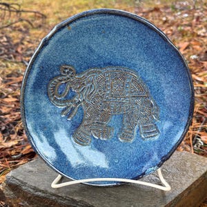 May include: A round, blue ceramic plate with a detailed embossed elephant design. The plate is displayed on a white metal stand, resting on a gray stone. The elephant is a light brown color.