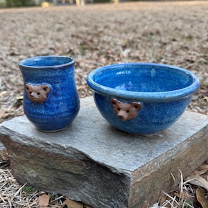 May include: Two handmade blue ceramic pieces: a small cup and a bowl. Each piece features a brown bear face applied to the exterior. The cup is cylindrical, and the bowl is round with a wide rim. Both are glazed with a glossy finish.