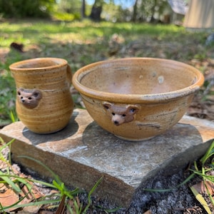 May include: A ceramic mug and a matching bowl with a speckled tan glaze. Each piece features a small, sculpted bear face attached to the exterior. The mug has a cylindrical shape with a handle, while the bowl has a wide, rounded rim.