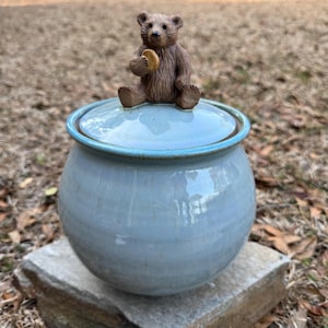 May include: A light blue ceramic jar with a lid, topped with a brown bear figurine holding a round object. The jar has a rounded shape and a light blue rim. The bear figurine is detailed with brown fur and sits atop the lid.