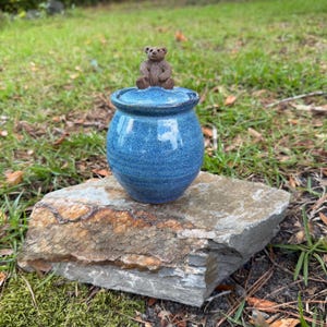 May include: A small, round, blue ceramic jar with a glossy, speckled finish. The lid features a sculpted, brown, seated bear figurine on top. The jar has a wide, rounded body that tapers slightly toward the base and a matching blue lid.