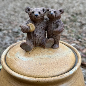 May include: A ceramic jar with a lid, topped with a brown bear figurine. One bear is seated and holding a small, round object, while another stands beside it. The jar and lid are tan with a speckled pattern, creating a rustic aesthetic.