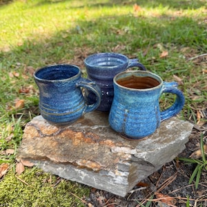 May include: Three ceramic mugs with handles, each featuring a unique blue glaze. One mug is a deep navy with horizontal ridges, another is a lighter mottled blue, and the third is a textured turquoise blue with a brown interior finish.