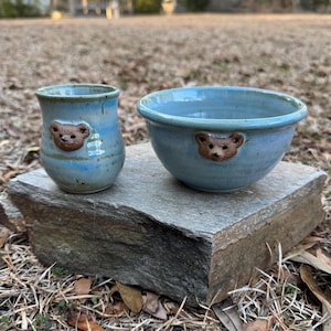 May include: A handmade ceramic set featuring a small tumbler and a bowl, both in a light blue glaze with brown bear face accents. The pieces are displayed on a stone surface, showcasing their unique, handcrafted quality.