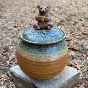 May include: A ceramic jar with a lid, featuring a brown bear figurine holding a biscuit. The jar has a round shape with a blue, tan, and brown color scheme. The jar is sitting on a stone.