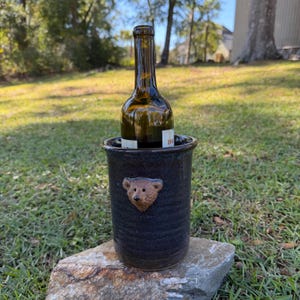 May include: A dark blue ceramic wine cooler with a textured surface and a sculpted brown bear face emblem on the front. An empty dark glass wine bottle is placed inside the cylindrical container, which rests on a rough, light-colored stone.