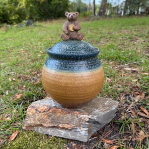 May include: A round ceramic jar with a lid featuring a sculpted brown bear holding a small circular object. The jar has a two-tone glaze, with a textured dark blue top section and a smooth, speckled tan bottom section, resting on a flat stone.