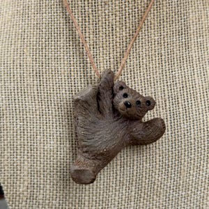 May include: A brown teddy bear pendant necklace. The bear is sculpted in a climbing pose, with a detailed texture. The necklace has a brown cord. The background is a textured, neutral-toned fabric.