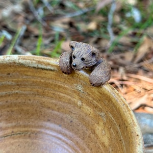 May include: A tan ceramic bowl with a textured, speckled finish featuring a small, brown, sculpted bear figurine attached to the rim. The bear is depicted with its front paws resting over the edge, looking inward toward the center of the bowl.