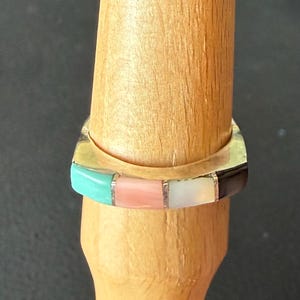 May include: A square-shaped ring with a silver band and a colorful inlay. The inlay features rectangular segments of turquoise, pink, white, and black stones. The ring is displayed on a wooden ring holder.