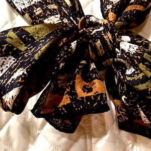 May include: A patterned scarf in shades of black, brown, white, and olive green. The scarf is knotted, showcasing the abstract design. The fabric appears to be silk or a similar lightweight material, suitable for various styling options.