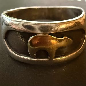 May include: A silver-toned ring with a bear silhouette in the center. The bear is gold-colored and set against a dark background. The ring has a thick band and a polished finish, suitable for everyday wear.