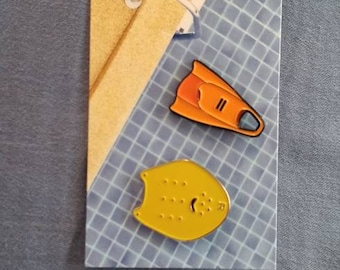 May include: Two enamel pins on a display card. One pin is a yellow swim paddle with holes, the other is an orange and black swim fin. The card has a blue and white grid pattern and a tan section.