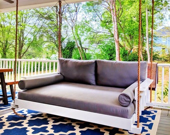 May include: White porch swing with gray cushions hanging from a wooden porch with a view of green trees and a blue and white patterned rug on the floor.