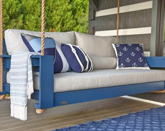 May include: A blue wooden swing with gray cushions and nautical-themed pillows. The swing is suspended by thick ropes and includes a blue and white striped throw blanket. The pillows feature anchor and rope designs.