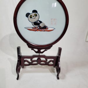 Puede incluir: Un expositor decorativo redondo con un panda en un barco. El panda es blanco y negro. El expositor tiene un marco y soporte de madera marrón oscuro.