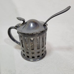 Puede incluir: Tarro de miel plateado con un tarro de vidrio y una cucharita. El tarro tiene un diseño de rejilla decorativo.