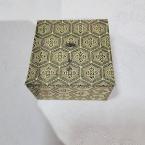 May include: A square box with a hinged lid featuring a repeating hexagonal pattern in shades of tan, olive green, and black. The surface has a textured fabric appearance with a symmetrical floral-like motif centered within each hexagon.