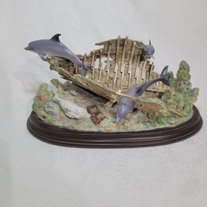 May include: A figurine of three dolphins swimming around a wooden shipwreck. The shipwreck is made of weathered wood and has a green moss-like growth on it. The dolphins are gray and white and have realistic details. The figurine is set on a brown oval base.