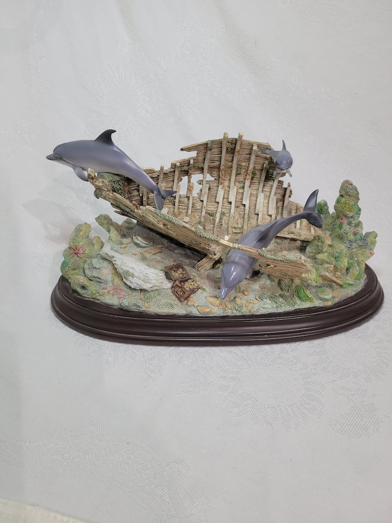 May include: A figurine of three dolphins swimming around a wooden shipwreck. The shipwreck is made of weathered wood and has a green moss-like growth on it. The dolphins are gray and white and have realistic details. The figurine is set on a brown oval base.