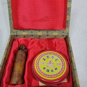 Peut inclure: Une boîte rouge avec un couvercle à motifs dorés est ouverte, révélant un tampon en bois avec un motif sculpté et une boîte ronde rouge et jaune d'encre. La boîte porte des caractères chinois et le mot "GUTA" imprimé dessus.