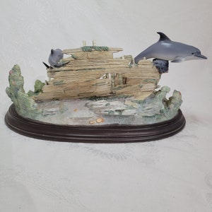 May include: A figurine of a dolphin jumping over a wooden shipwreck with coral and seaweed surrounding it. The figurine is on a brown oval base.