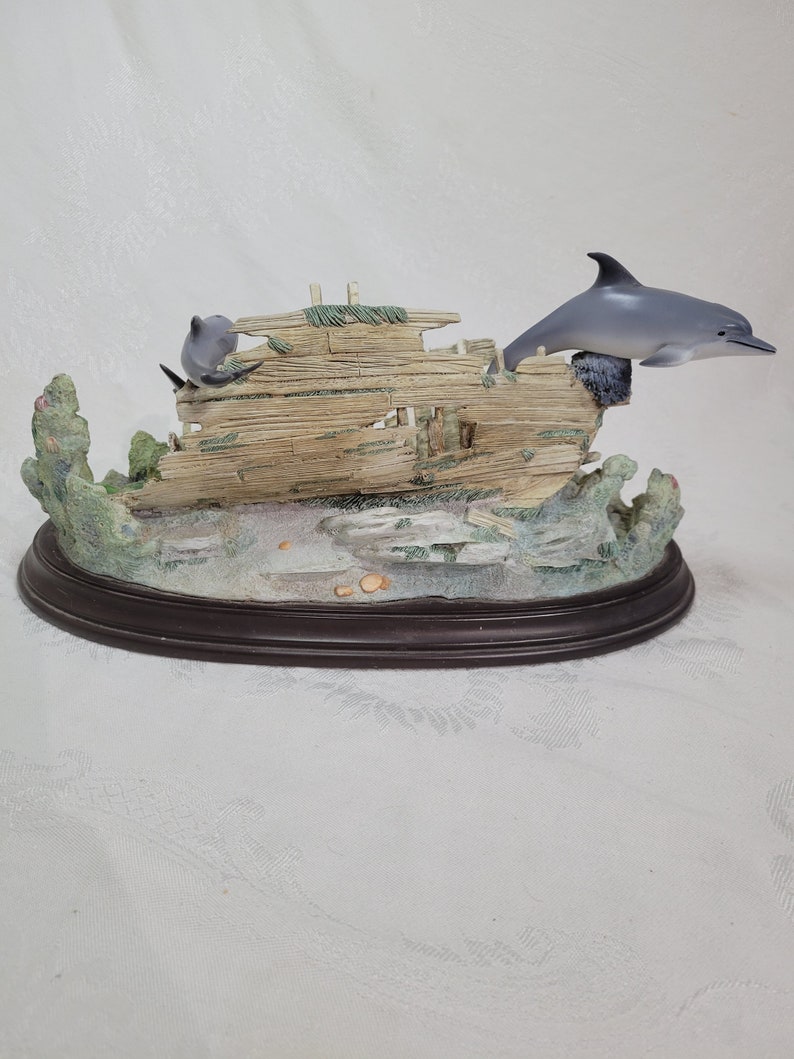 May include: A figurine of a dolphin jumping over a wooden shipwreck with coral and seaweed surrounding it. The figurine is on a brown oval base.