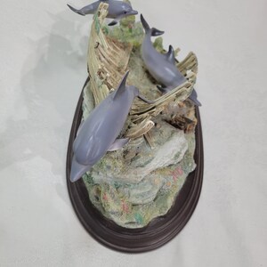 May include: A figurine of three gray dolphins jumping out of a broken wooden boat. The figurine is set on a brown oval base with a rocky, green, and coral-colored base.