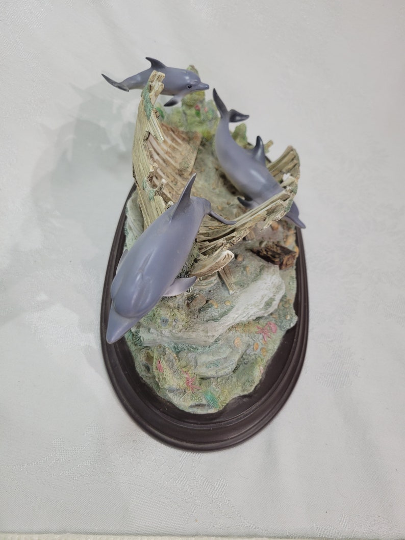 May include: A figurine of three gray dolphins jumping out of a broken wooden boat. The figurine is set on a brown oval base with a rocky, green, and coral-colored base.