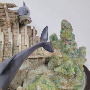 May include: A gray and white dolphin figurine leaping out of a wooden shipwreck, surrounded by green and brown coral and seaweed.