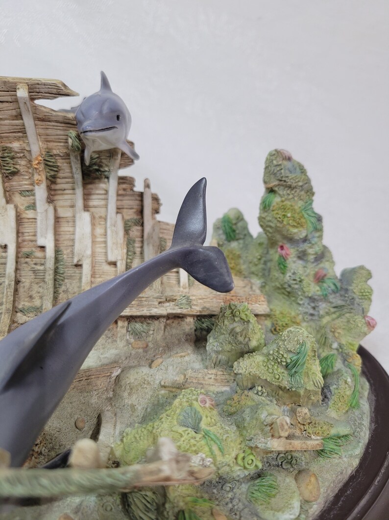 May include: A gray and white dolphin figurine leaping out of a wooden shipwreck, surrounded by green and brown coral and seaweed.