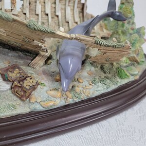 May include: A ceramic figurine of a shipwreck with three dolphins swimming around it. The shipwreck is made of wood and has a weathered look. The dolphins are painted in shades of gray and white. The figurine is set on a brown oval base.