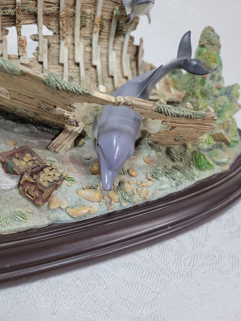 May include: A ceramic figurine of a shipwreck with three dolphins swimming around it. The shipwreck is made of wood and has a weathered look. The dolphins are painted in shades of gray and white. The figurine is set on a brown oval base.
