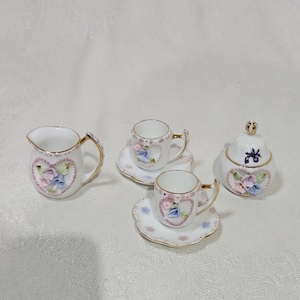 May include: A miniature porcelain tea set featuring a creamer, a sugar bowl with a lid, and two cups with matching saucers. Each piece is white with gold trim, decorated with raised pink and blue floral heart motifs and small painted flowers.