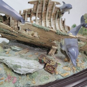 May include: A figurine of a shipwreck with three dolphins swimming around it. The shipwreck is made of wood and has a treasure chest with gold coins inside. The base of the figurine is brown.