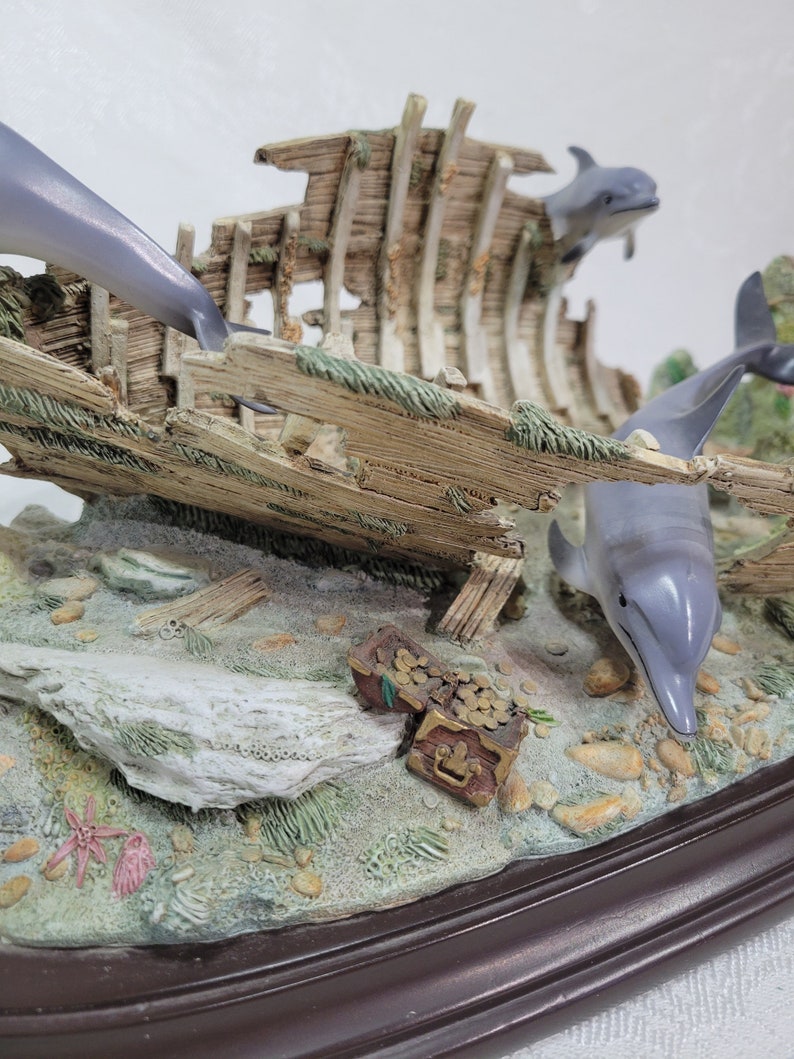 May include: A figurine of a shipwreck with three dolphins swimming around it. The shipwreck is made of wood and has a treasure chest with gold coins inside. The base of the figurine is brown.