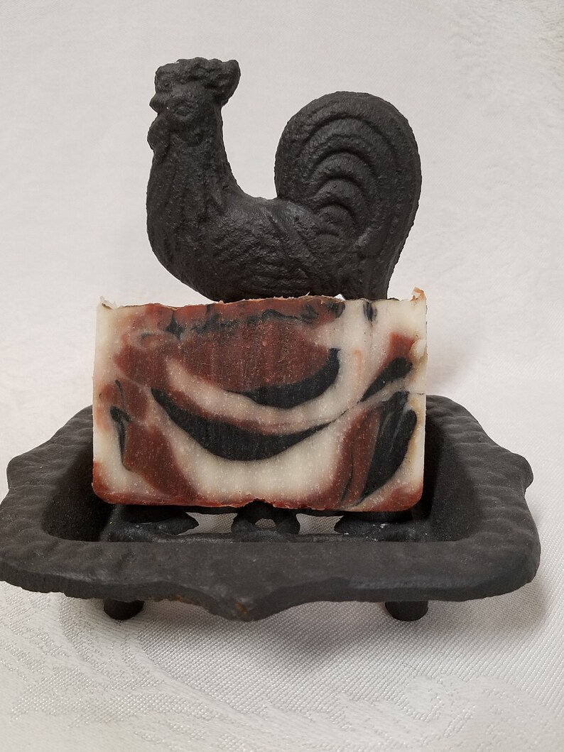 May include: A bar of handmade soap with brown and white swirls sits on a black cast iron soap dish with a black rooster figurine on top.