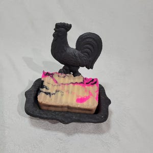 May include: A black rooster soap dish with a pink and white swirl soap bar. The soap dish is made of cast iron and has a decorative design.