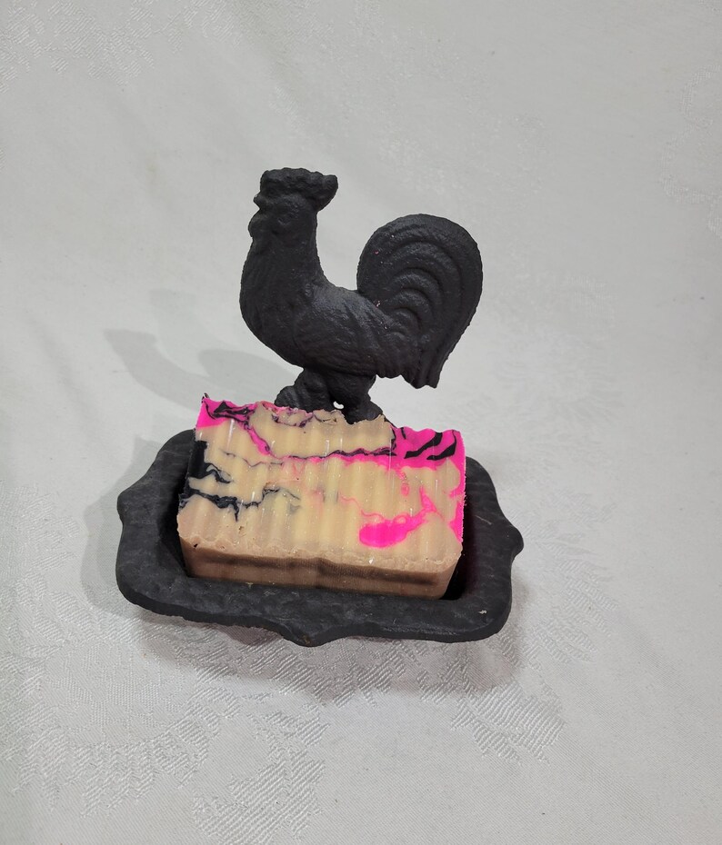 May include: A black rooster soap dish with a pink and white swirl soap bar. The soap dish is made of cast iron and has a decorative design.