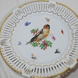Puede incluir: Plato de porcelana blanca con borde dorado y diseño calado intrincado. El plato presenta una ilustración detallada de un pájaro posado en una rama floreciente, rodeado de mariposas e insectos. Una pieza decorativa.