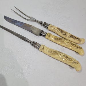 Puede incluir: Juego de trinchar antiguo con cuchillo, tenedor y afilador. Las hojas muestran desgaste, mientras que los mangos son de color crema con diseños de hojas ornamentados. El juego incluye un cuchillo para trinchar, un tenedor para trinchar y un afilador.