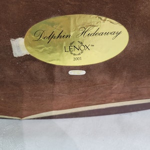 May include: A gold oval sticker with the text "Dolphin Hideaway" and "LENOX" in a stylized font. The sticker also includes the year "2001".