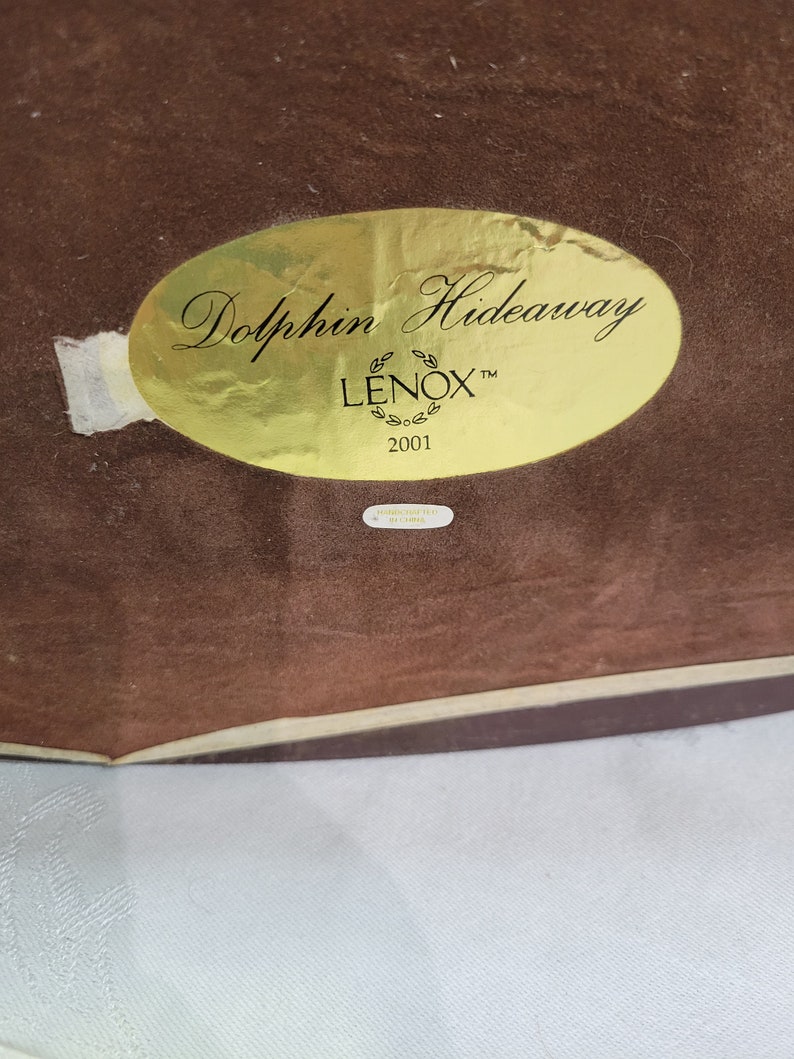 May include: A gold oval sticker with the text "Dolphin Hideaway" and "LENOX" in a stylized font. The sticker also includes the year "2001".