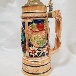 May include: A ceramic beer stein featuring a detailed scene of a house, trees, and flowers. The stein has a hinged lid with a metal handle and a brown, sculpted handle.
