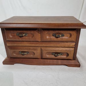 Puede incluir: Una caja de joyería de madera con cuatro cajones. La caja es de color marrón cálido con tiradores de metal ornamentados. La parte superior de la caja es plana y la base está ligeramente elevada.