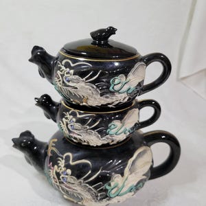 May include: A stacked black ceramic tea set with a teapot and two cups. The set features dragon and floral designs in white, silver, and teal. The teapot has a bird-shaped handle and a bird-shaped lid finial.