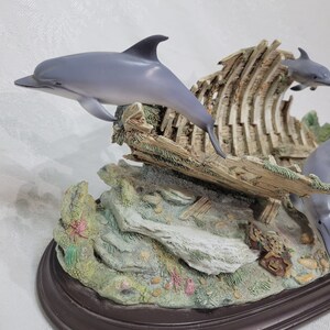 May include: A figurine of three dolphins jumping out of the water near a shipwreck. The dolphins are grey and white, and the shipwreck is made of wood. The figurine is set on a base that is covered in green moss and has a treasure chest.